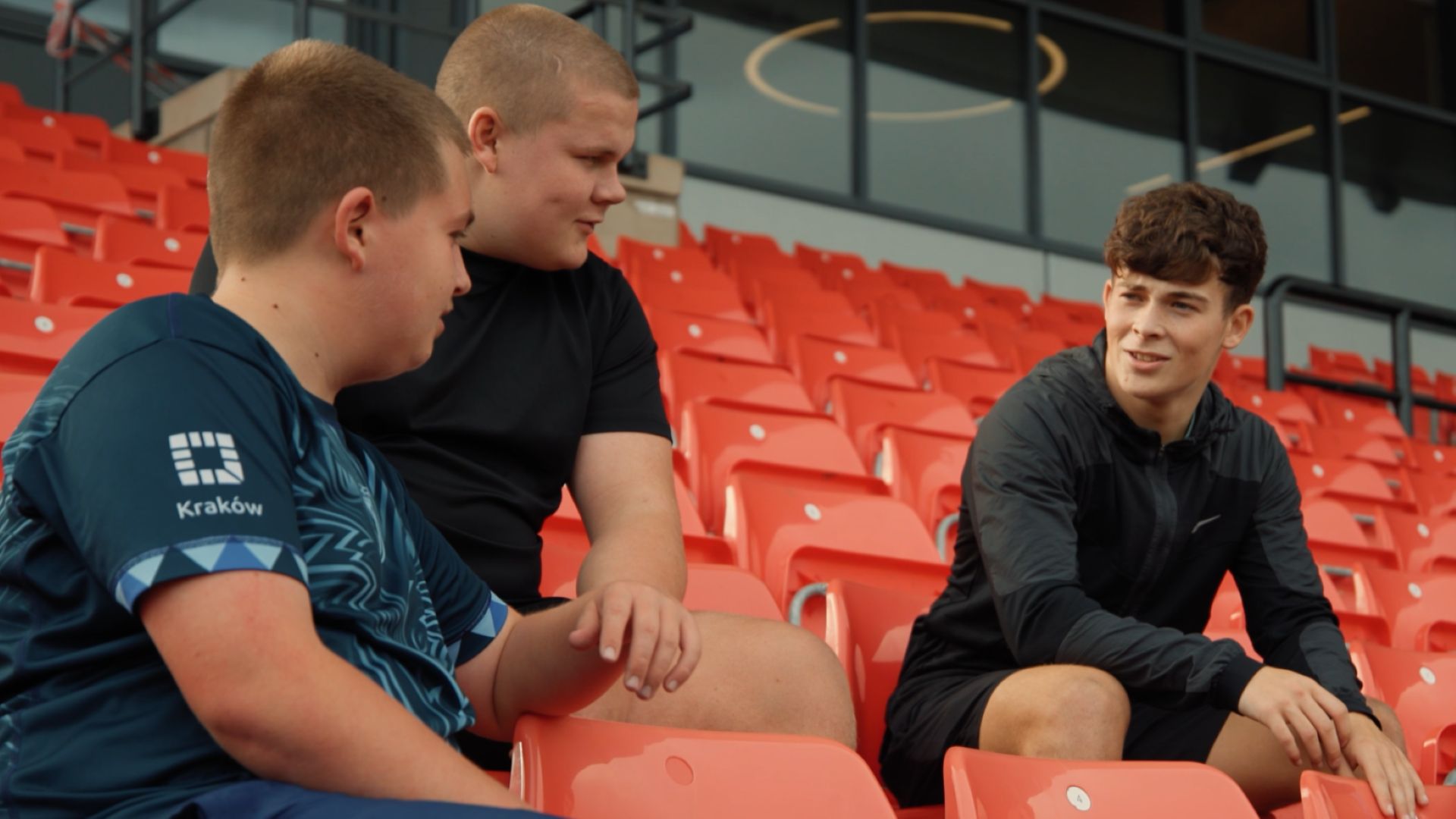 3 young people sat in the stands of a stadium on red seating