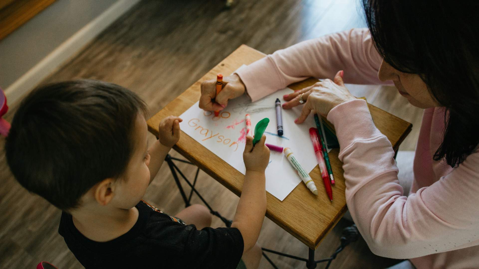 child with crayons writing name on paper with teacher kneeling next to them.