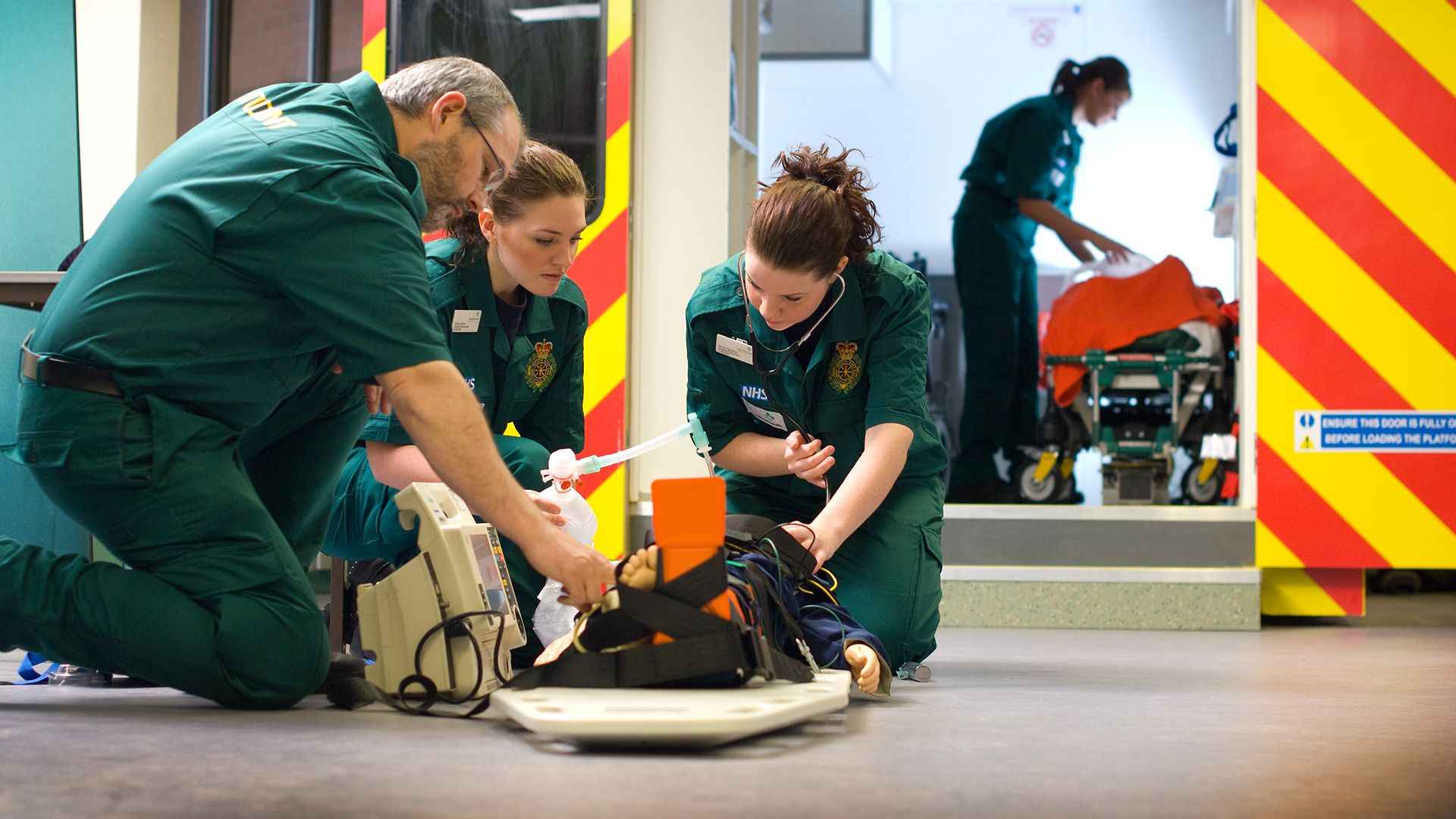 Paramedics treat a patient on a stretcher beside an ambulance, using monitoring and breathing equipment during a simulated emergency response.