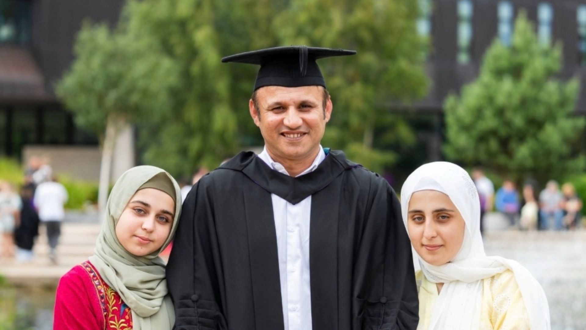 Graduate standing outside with two daughters beside him wearing hijab and sunshine.