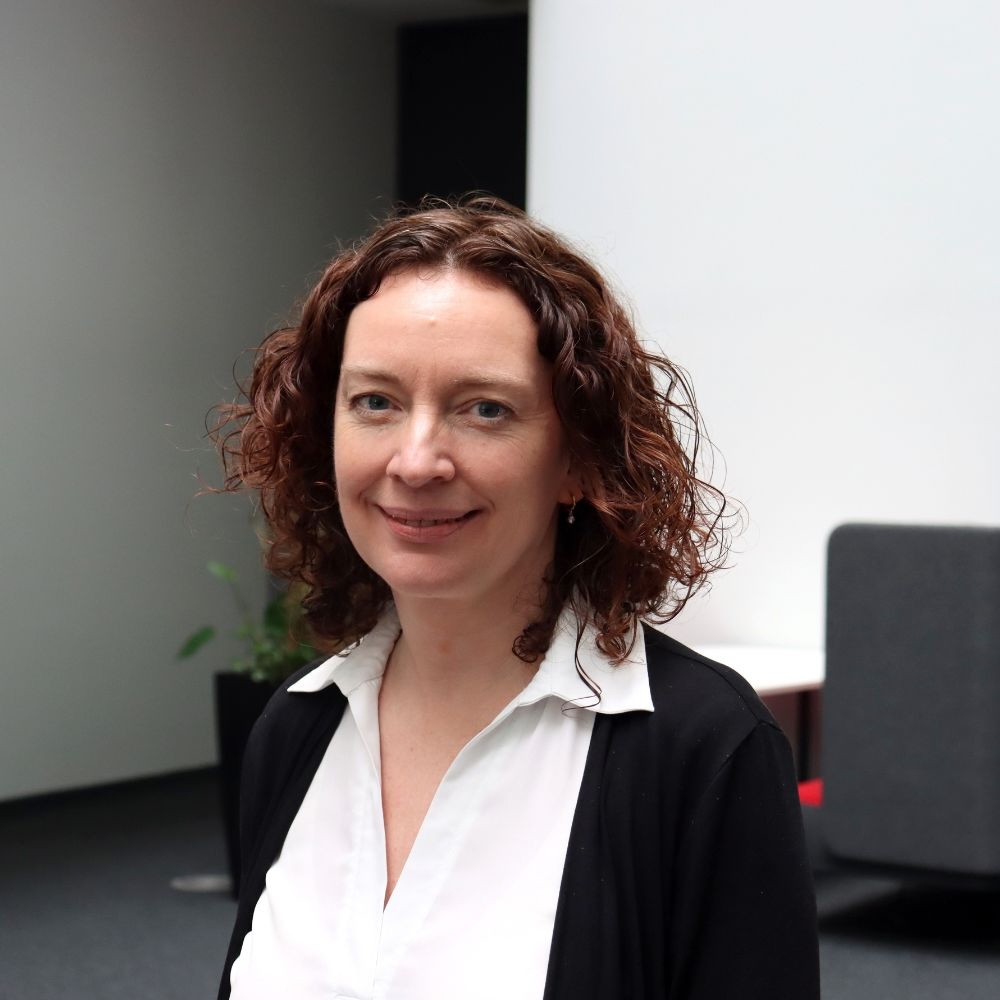 profile photo of Sarah looking straight at the camera smiling with a white shirt, black blazer and curly redish hair. 